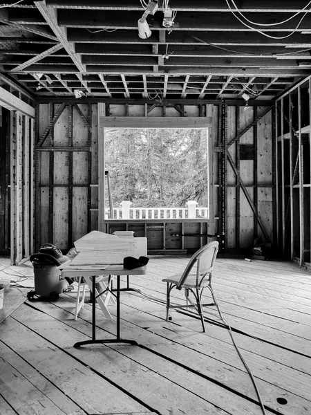 Black and white image of a partially constructed room with exposed wooden beams, a table, chairs, and a large window overlooking trees.